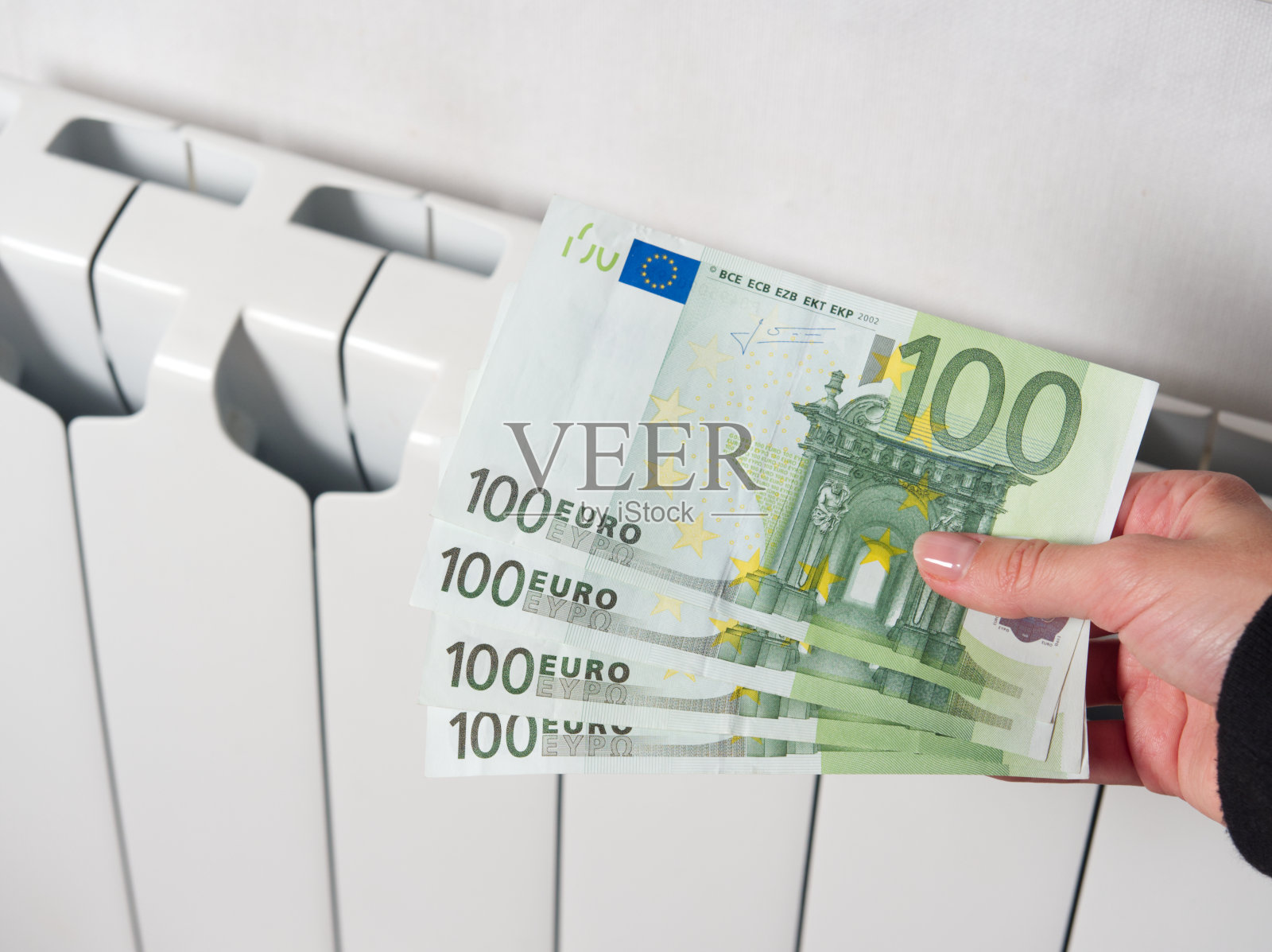 一只手拿着一百欧元钞票的特写。背景中，中央供暖电池是白色的。欧洲的高支付概念。水电费增加。照片摄影图片