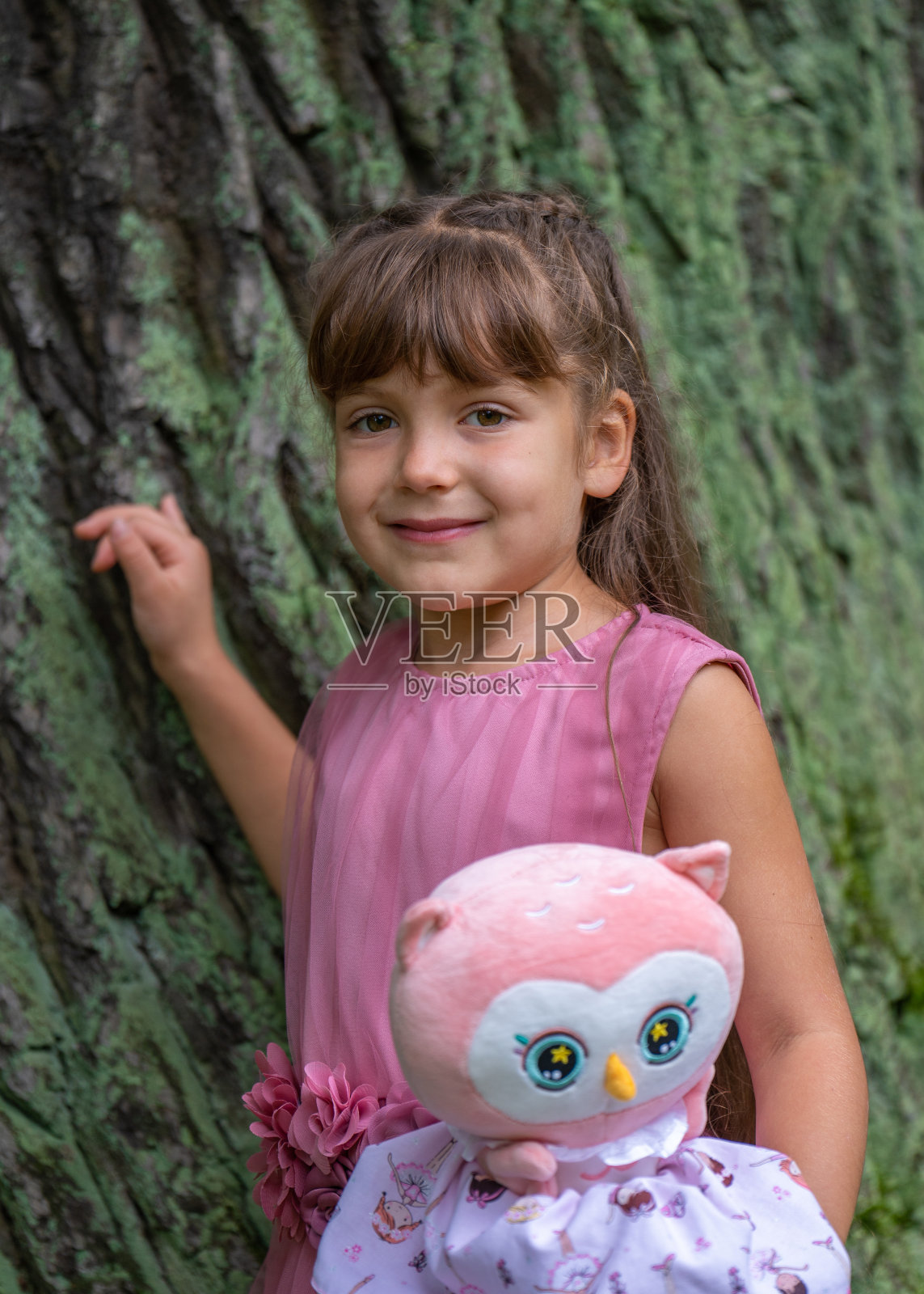 一个可爱的小女孩站在一棵大树旁边，高兴地看着镜头，手里拿着一个玩具照片摄影图片