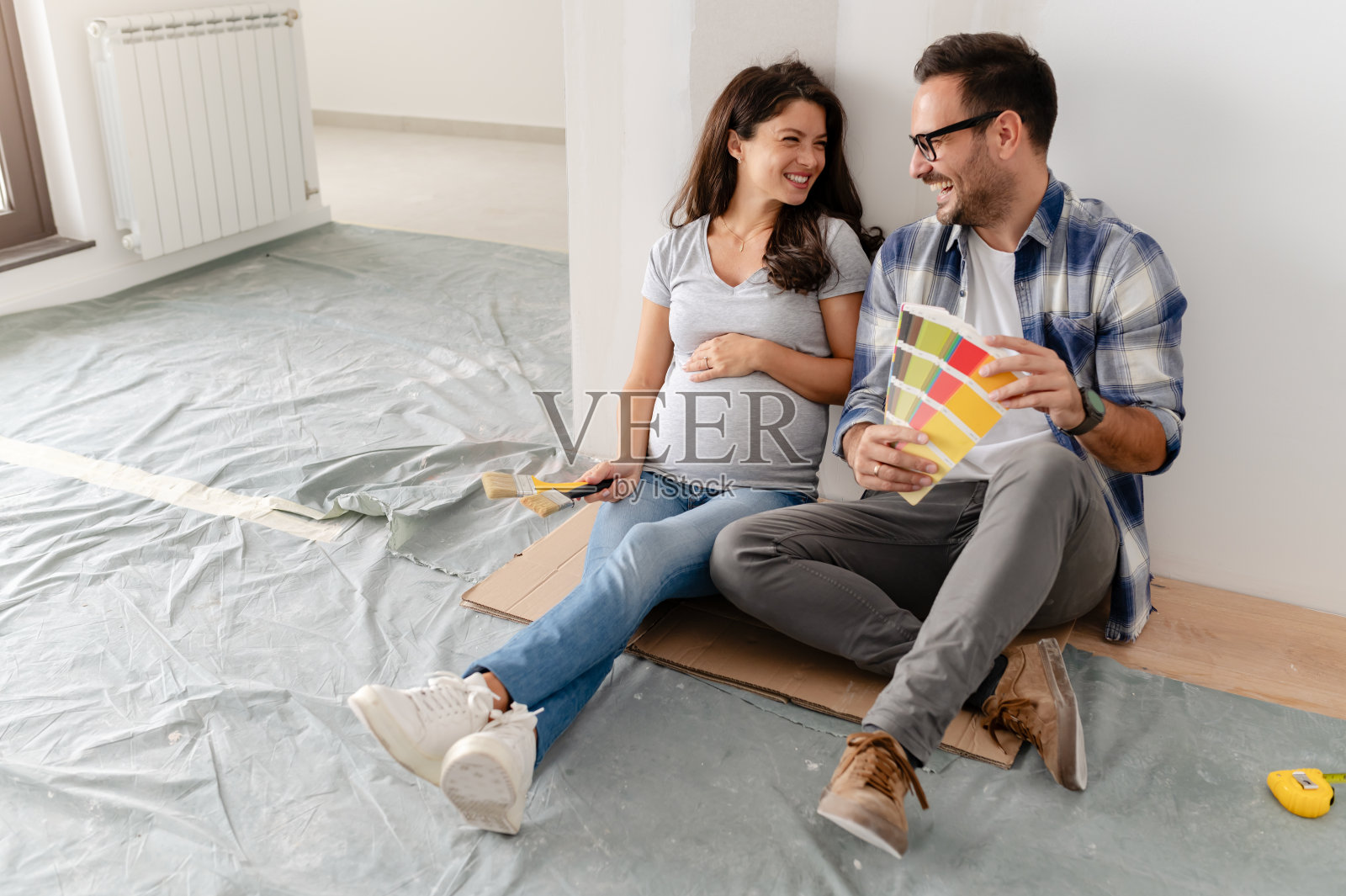 一对年轻、美丽、快乐的夫妇，包括一个男人和他怀孕的妻子，坐在一间空房子的地板上。他们正在挑选装饰新家的颜色照片摄影图片