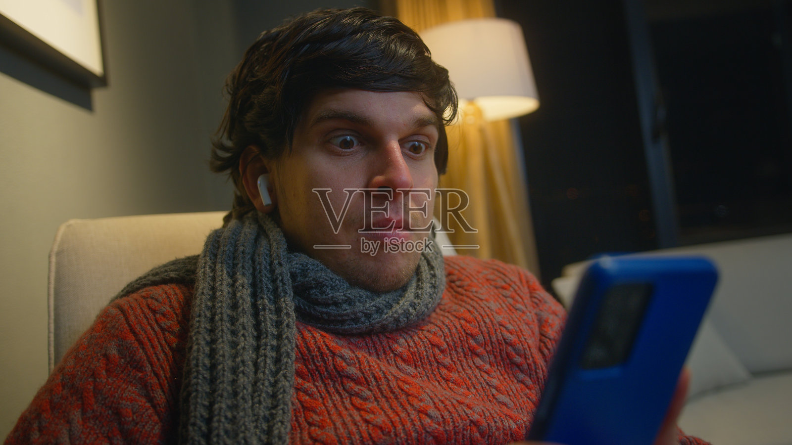 男人戴着无线入耳式耳机，坐在客厅的扶手椅上，用智能手机浏览互联网，看视频，使用社交网络照片摄影图片