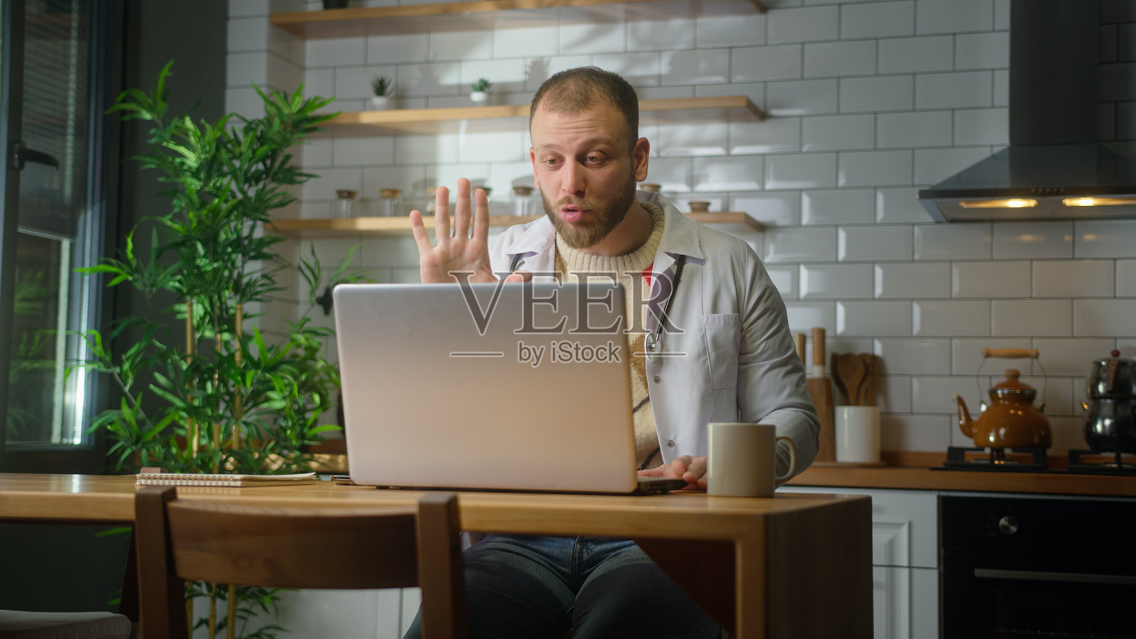 专业男医生穿着白色制服，戴着听诊器进行会议视频通话，通过笔记本电脑的网络摄像头与患者在线聊天。远程医疗咨询照片摄影图片