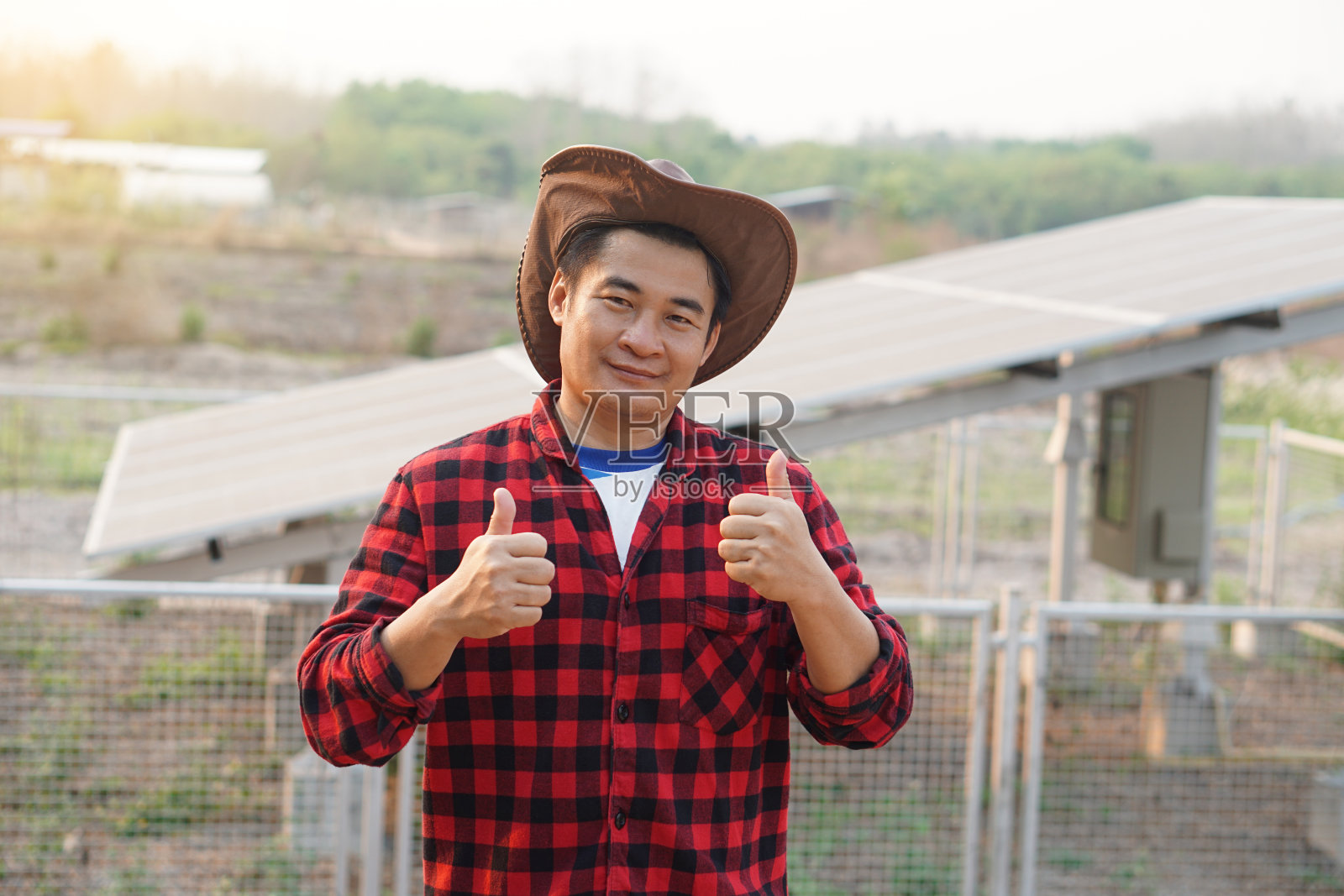 英俊的亚洲男子戴着帽子，穿着红色格子衬衫，站在农业用地的太阳能电池板旁。照片摄影图片