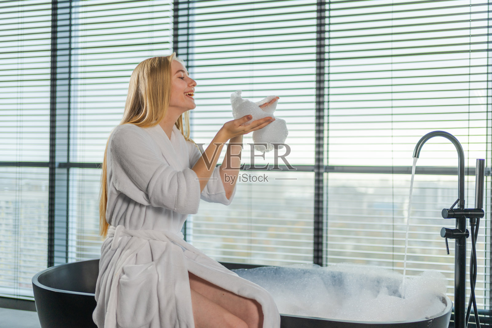 水疗中心放松。穿着浴袍的年轻女子坐在浴池里，在浴缸里吹泡沫。女孩在家里的浴室里放松。女人在浴室里度过了一上午。美容护理身体护肤理念。照片摄影图片