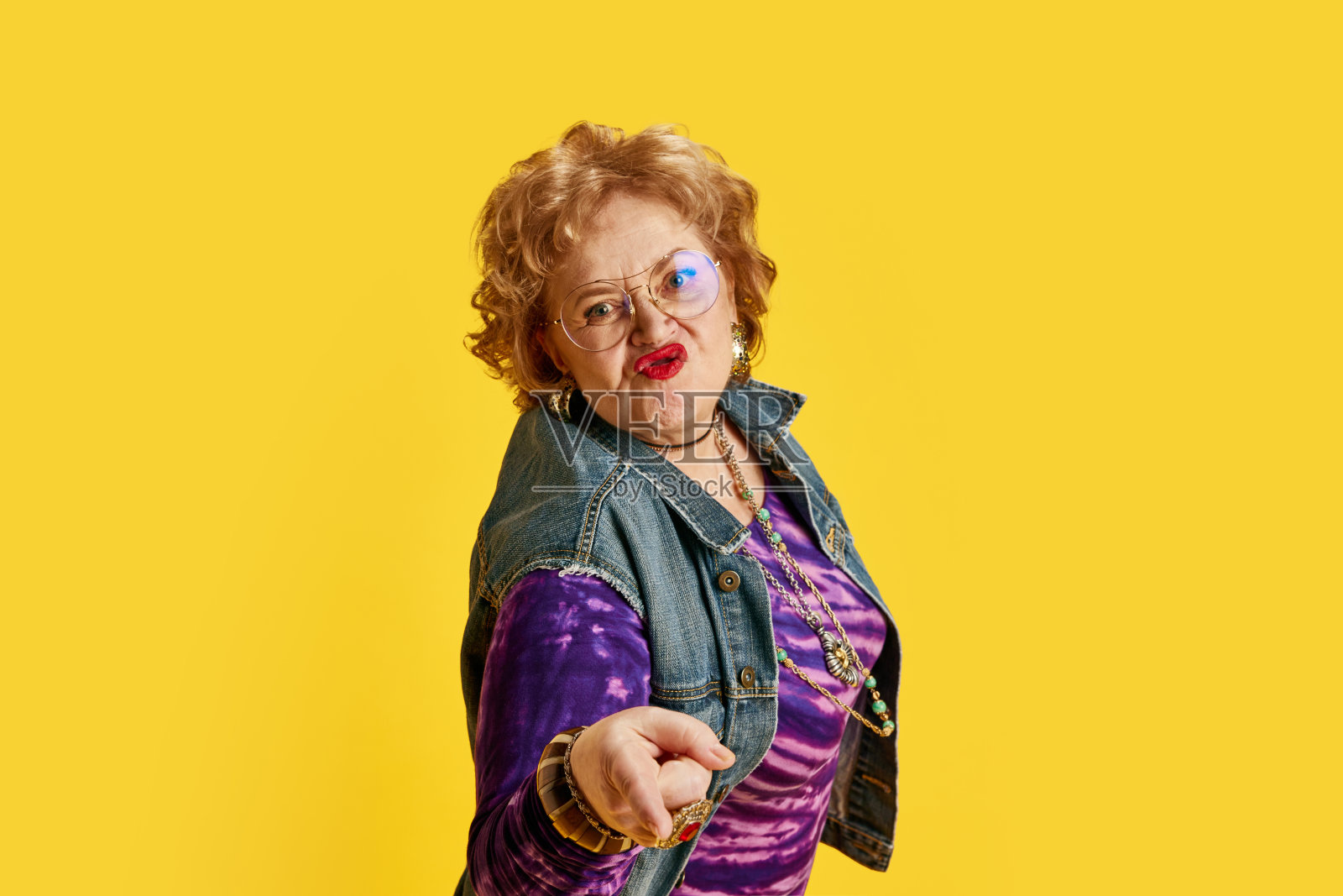 一位戴着圆眼镜抿着嘴唇的年长女子穿着牛仔夹克，紫色t恤上镶嵌着黄色背景的黄金首饰。照片摄影图片