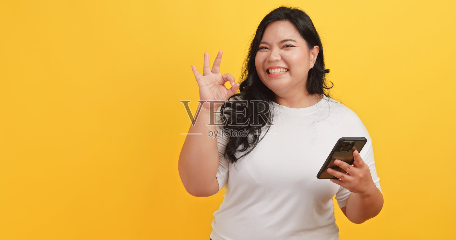 一位穿着白色t恤的胖乎乎的亚洲女性看到黄色背景的手机上有一条信息，兴奋而高兴照片摄影图片
