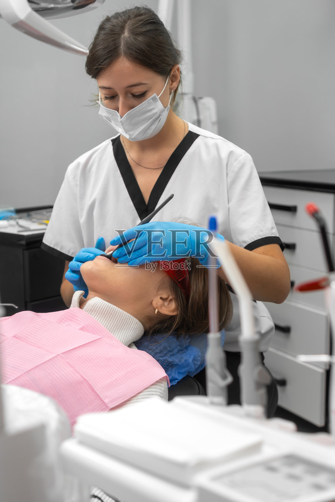 女牙医穿着制服，戴着医用口罩，在牙科诊所检查病人的牙齿。漂亮的病人躺在牙科椅上接受牙医的牙科治疗。垂直图照片摄影图片