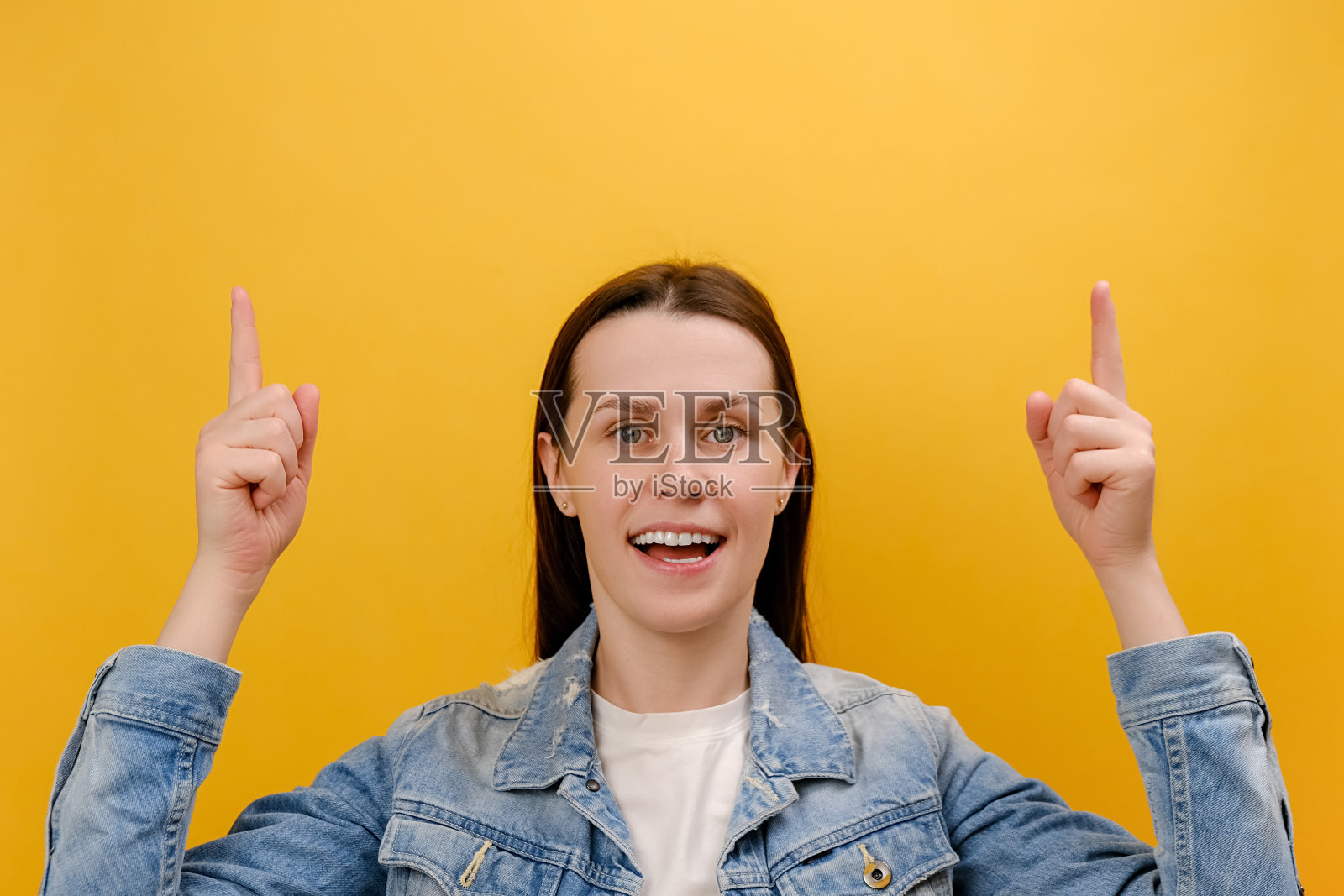 一个惊喜的年轻女子的肖像，她指着上面，带着震惊的表情，展示了商业广告的复制空间，穿着牛仔夹克，孤立在工作室的黄色背景墙上照片摄影图片