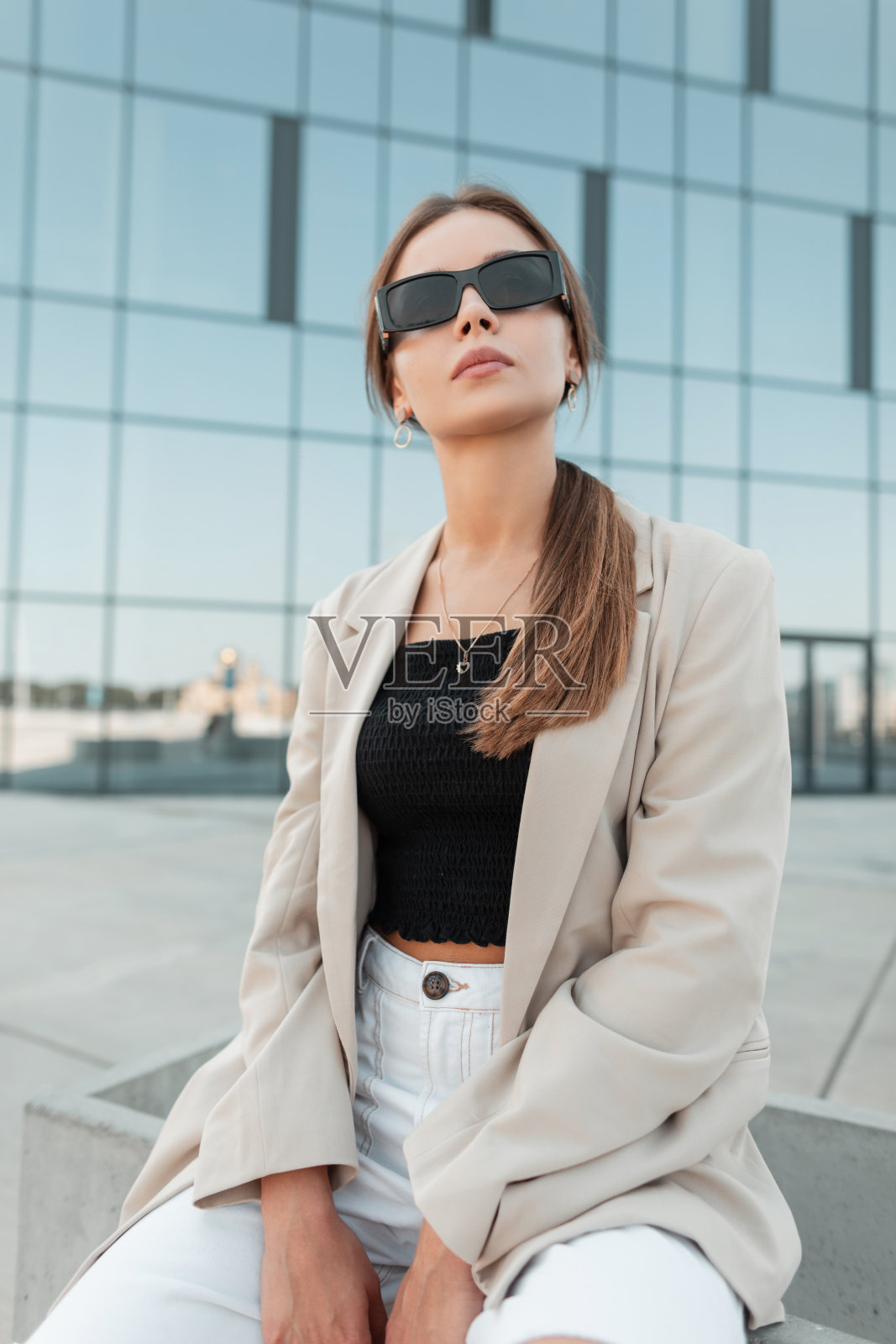 戴着墨镜的漂亮时尚女孩坐在城市里，背景是一座蓝色的玻璃建筑照片摄影图片