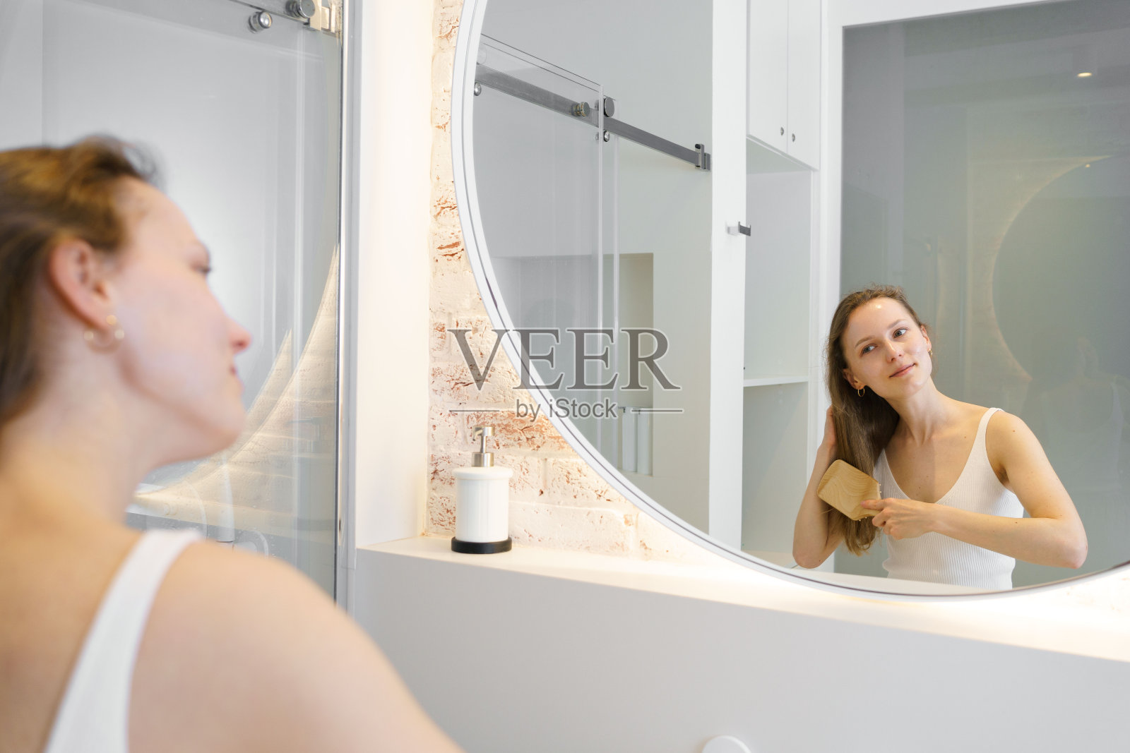 一个年轻的棕色头发的女人一边看着浴室里的镜子，一边用一把木梳子梳理着她闪亮的头发。护发和化妆品的概念。照片摄影图片