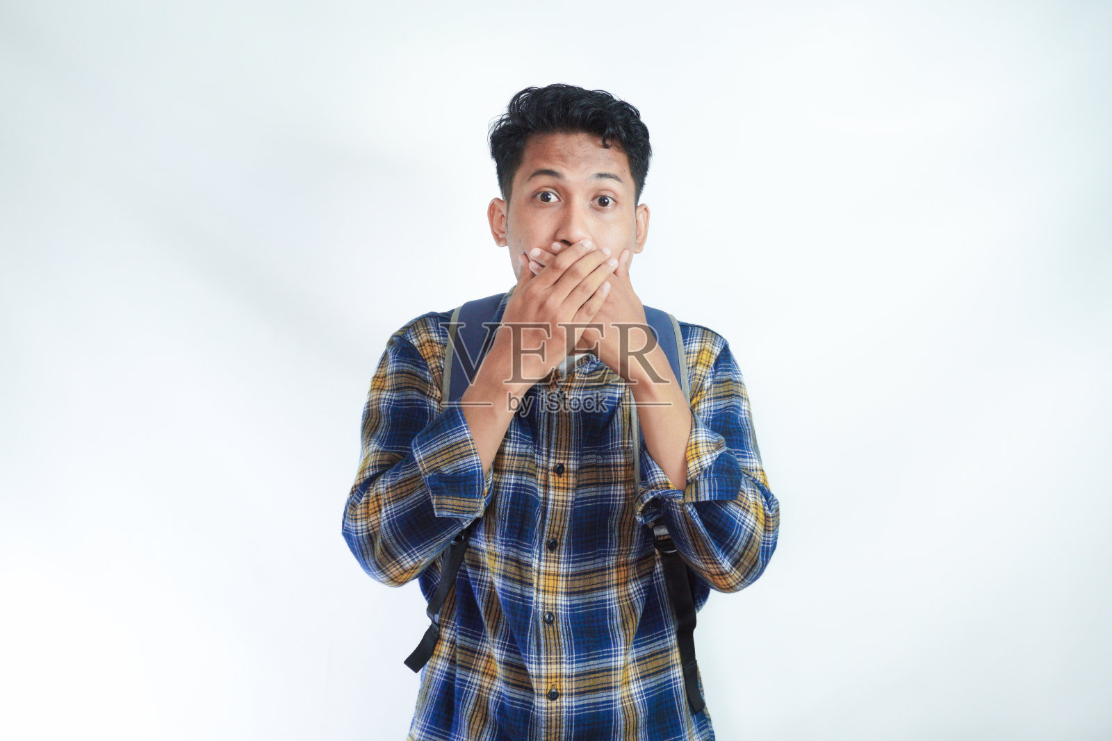 震惊惊讶担心年轻的亚洲学生男子20多岁，穿着休闲服，背着背包，用手捂住嘴，看着白色背景的相机。高中大学学院照片摄影图片