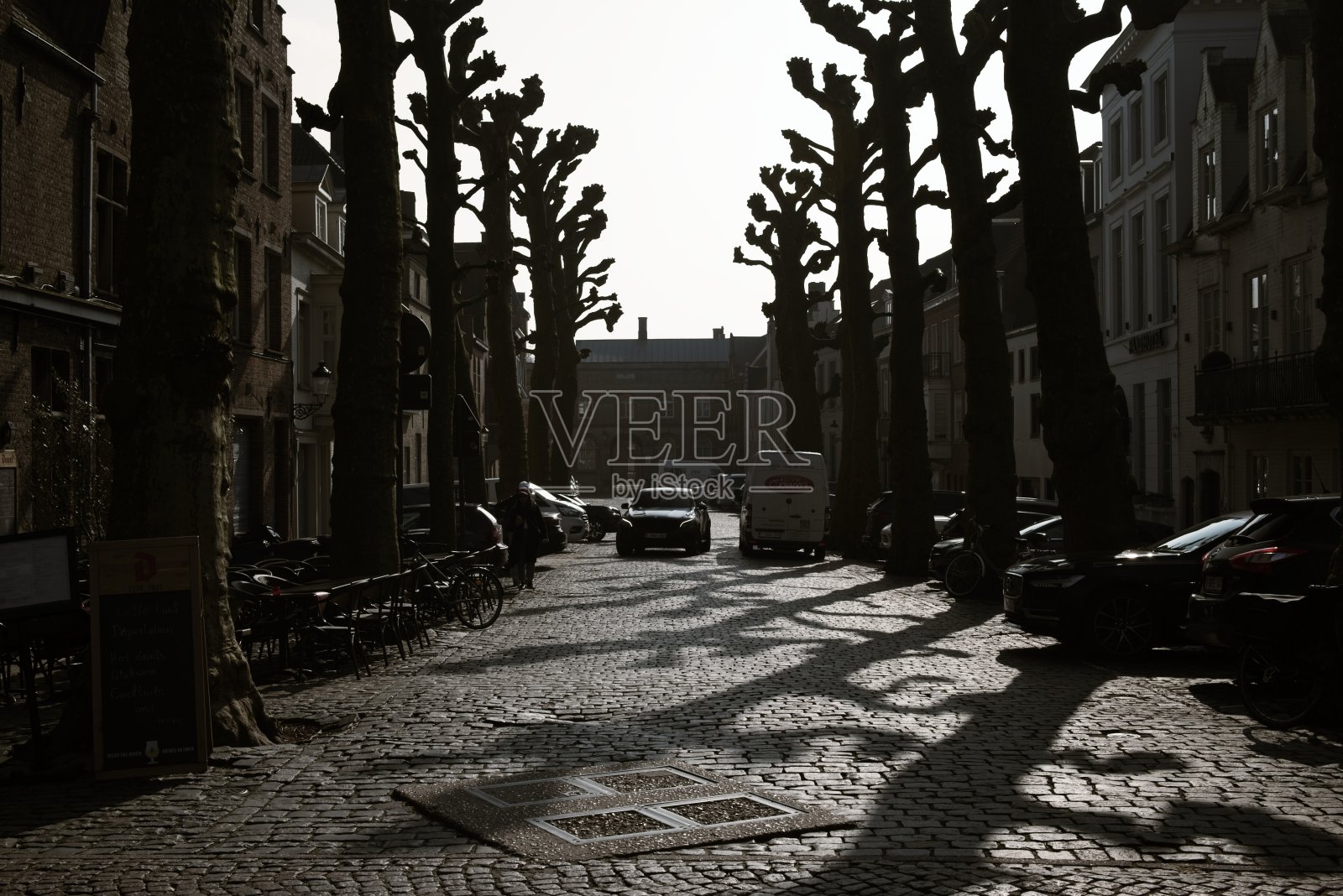 在一个古雅的欧洲小镇，黄昏时分，鹅卵石铺成的街道上，修剪过的树木投下了剪影，汽车停在路边。照片摄影图片