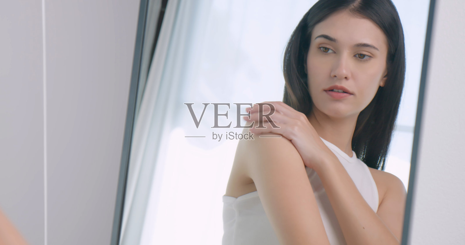 美丽的年轻女子站在浴室的镜子前，在身上涂抹润肤霜。照片摄影图片