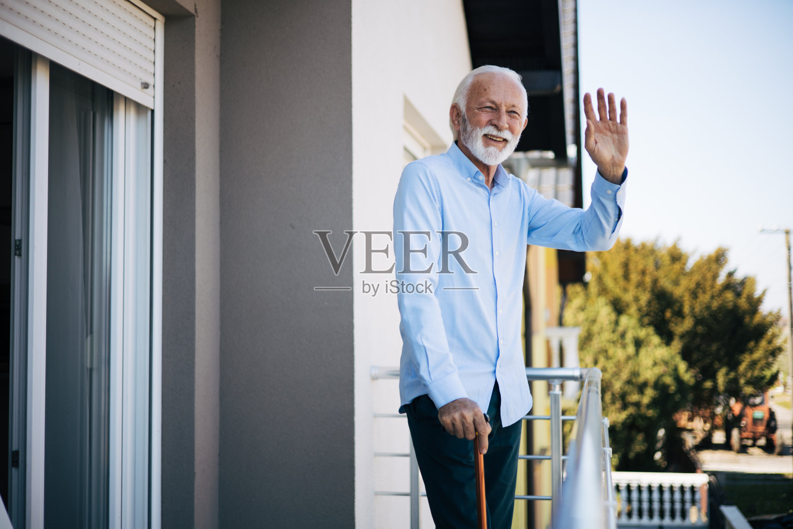 一位老人站在自家的露台上挥手，拄着拐杖走路。照片摄影图片