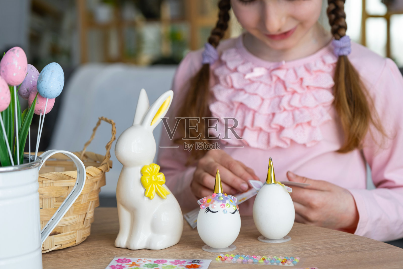 一个长着粉色兔子耳朵的可爱女孩做了一个复活节工艺品——用水钻、角、花把一个鸡蛋装饰成独角兽的形状，在一个有植物的房子里。照片摄影图片