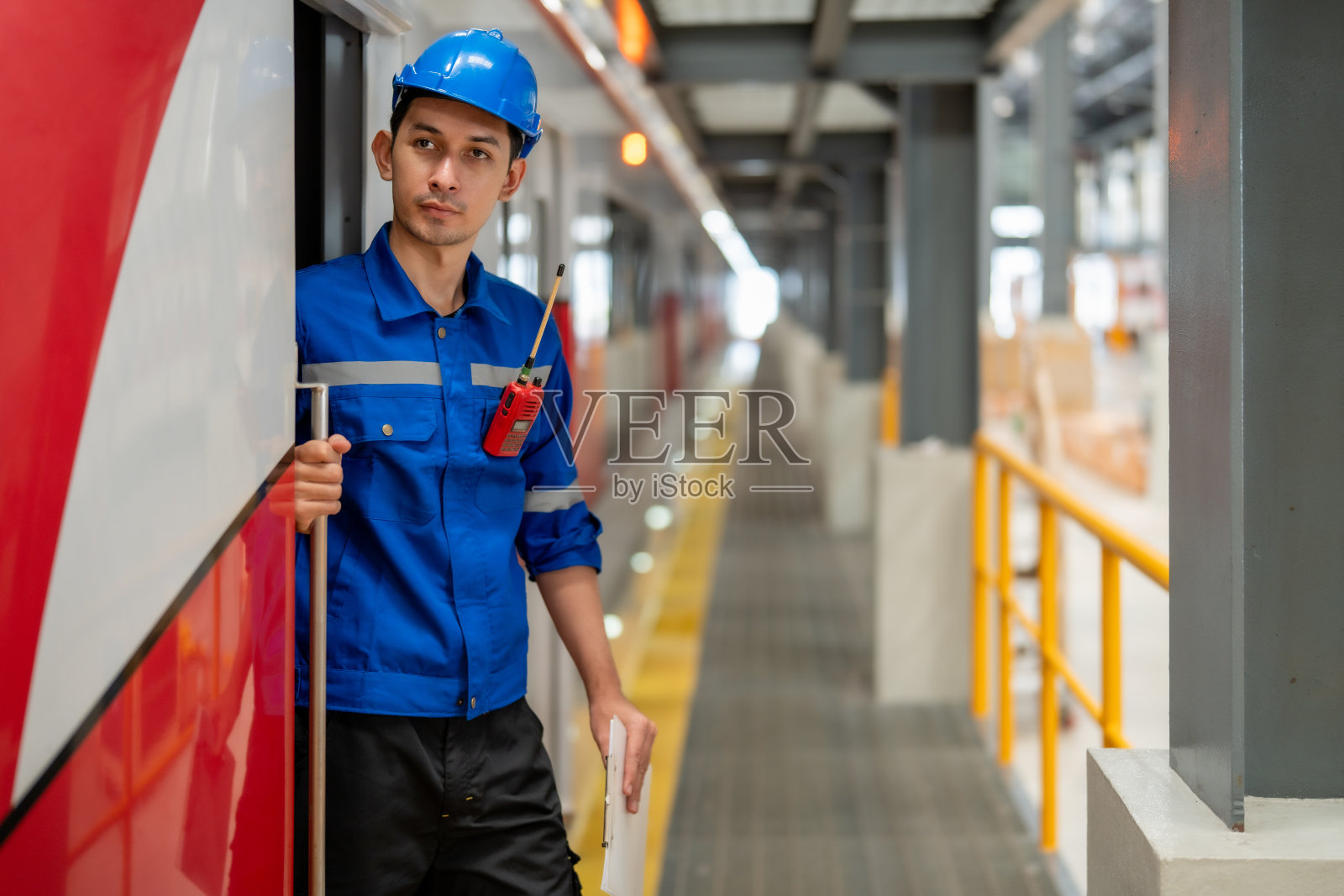 一名身穿蓝色制服、头戴防护头盔的列车员站在车门旁，手持对讲机和文件。照片摄影图片