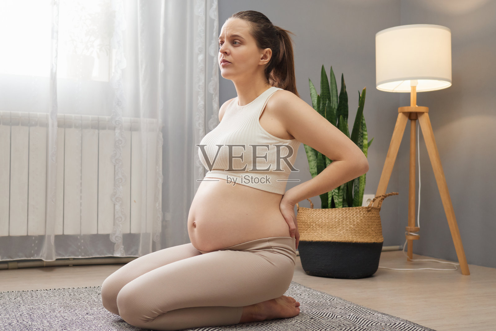 疲惫的孕妇穿着米色的衣服，腰酸背痛，摸着疼痛的背部，感觉不舒服，坐在客厅的地板上，感觉疲劳照片摄影图片