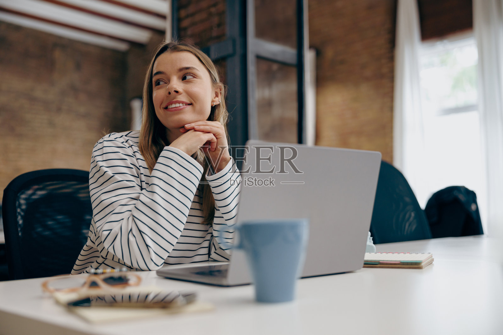 年轻漂亮的女经理坐在办公室的桌子上，一边用笔记本电脑工作，一边看向别处照片摄影图片