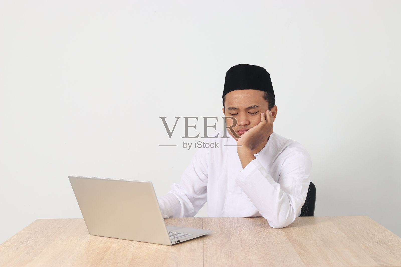 疲惫的亚洲穆斯林男子的肖像，穿着科科衬衫，戴着无边帽，在斋月斋戒期间工作，感到困倦，打着哈欠，用手捂住嘴。白色背景上的孤立图像照片摄影图片