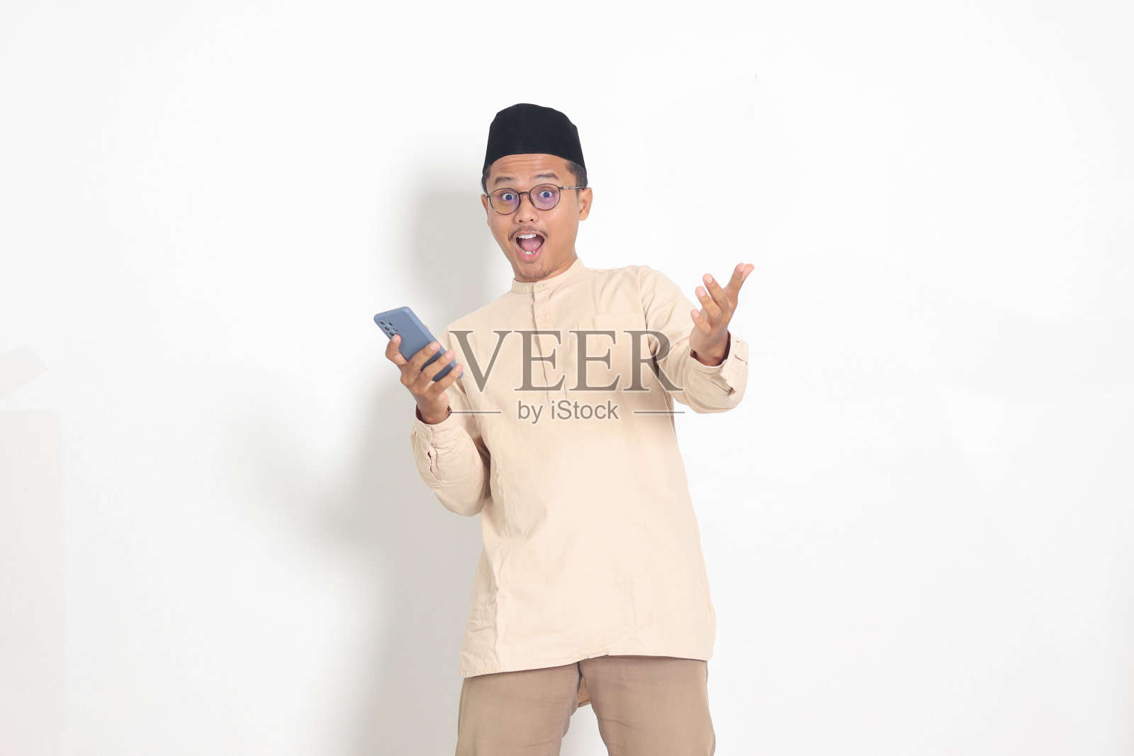 吃惊的亚洲穆斯林男子的肖像，身穿科科衬衫，戴着无边便帽，手里拿着手机，脸上露出震惊的表情。广告和社交媒体概念。白色背景上的孤立图像照片摄影图片