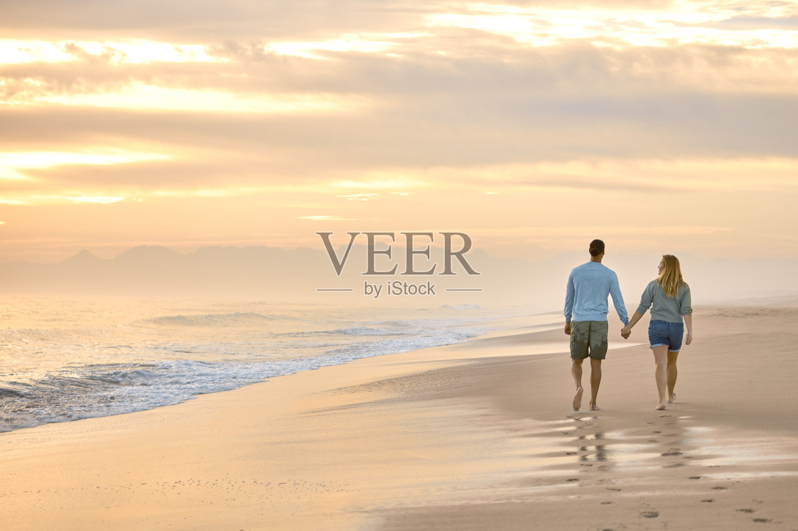 一对穿着休闲装度假的夫妇的后视图，在黎明时分牵着手沿着海滩散步照片摄影图片