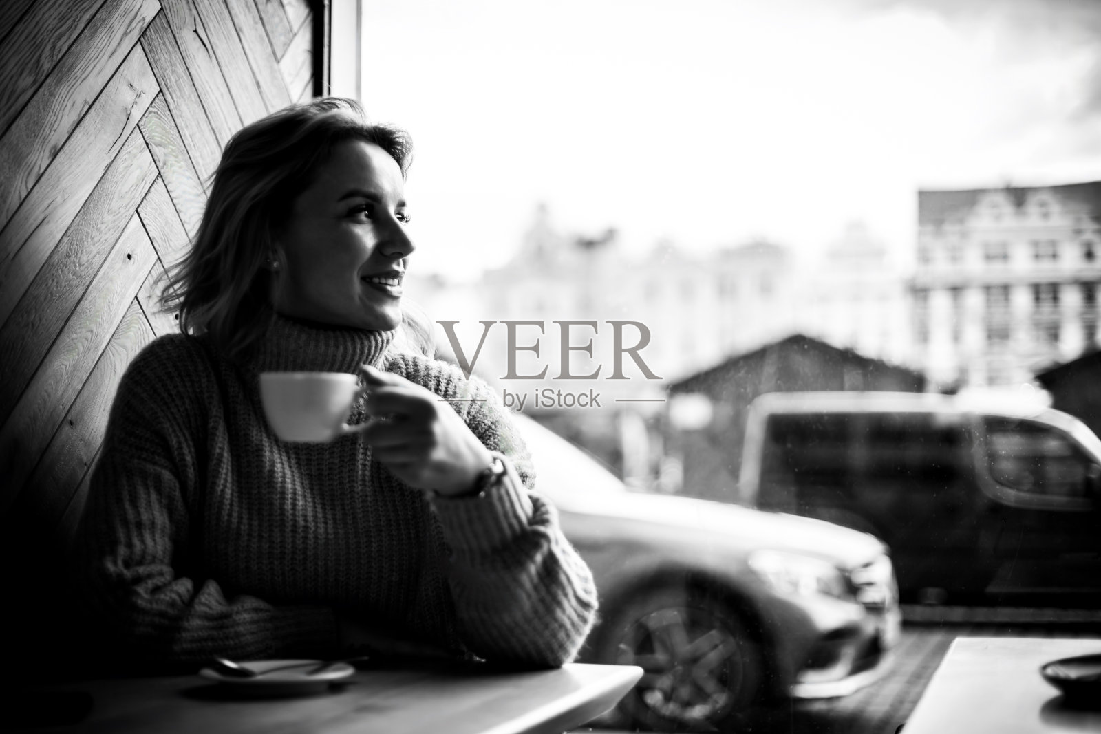 一位女士在城市咖啡馆靠窗享用咖啡。一个心满意足的女人坐在咖啡桌旁，手里端着一杯咖啡，微笑着凝视着窗外。外面的城市景象是一辆停着的汽车。照片摄影图片