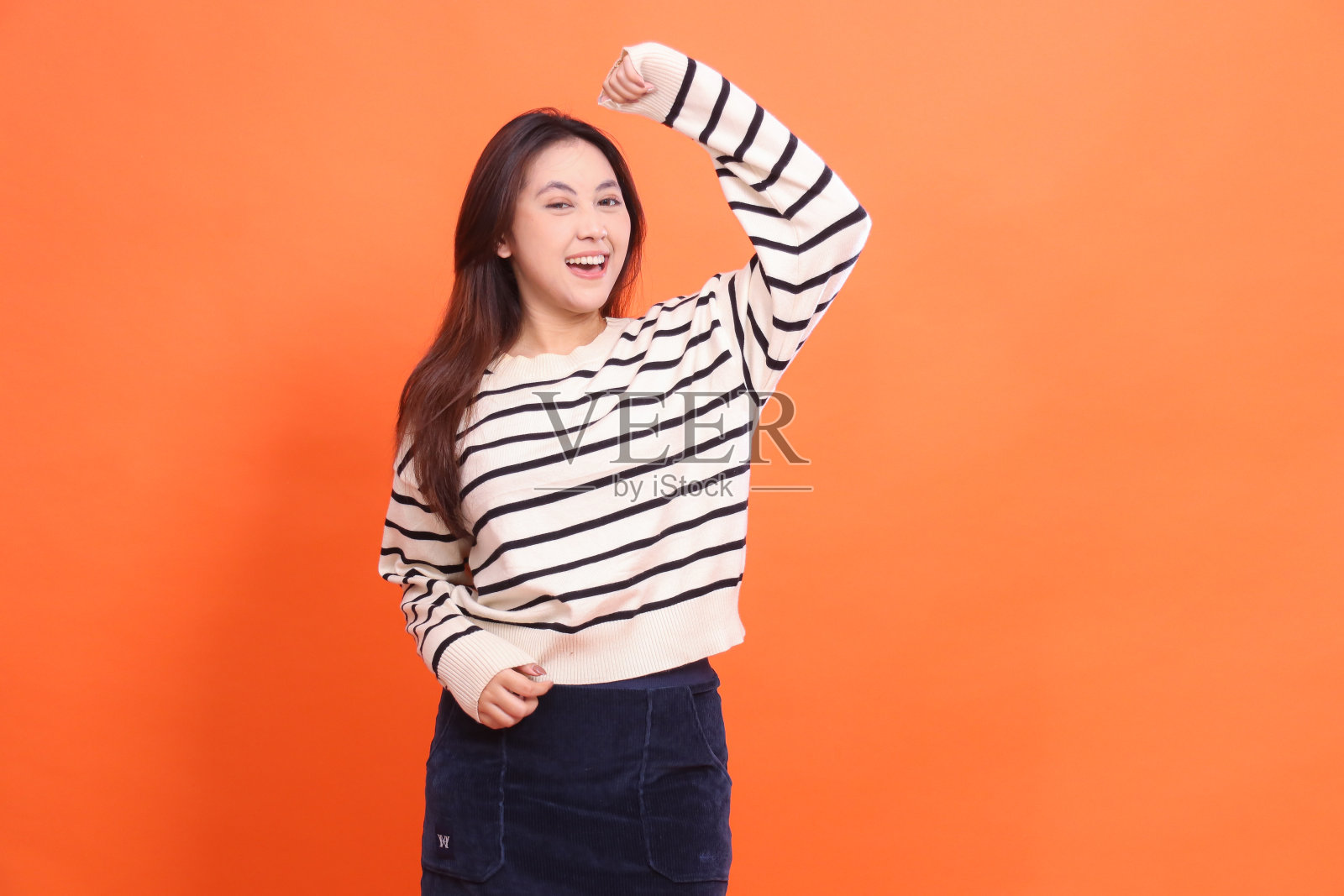 美丽、开朗、自信的印尼女子站在那里，紧攥着拳头，看着橙色背景下的镜头，穿着白色毛衣照片摄影图片