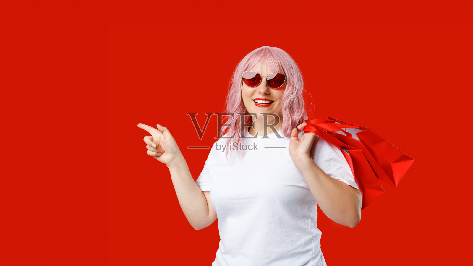 戴着墨镜的粉色头发的漂亮开朗的女人拿着购物袋，在红色背景的复制空间里伸出手指。特殊报价照片摄影图片