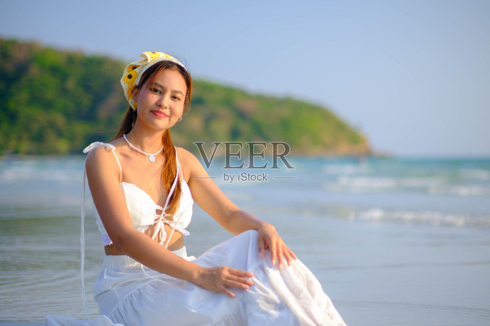 穿着白色连衣裙的年轻漂亮的亚洲女子坐在海边的水里，看起来很开心。照片摄影图片