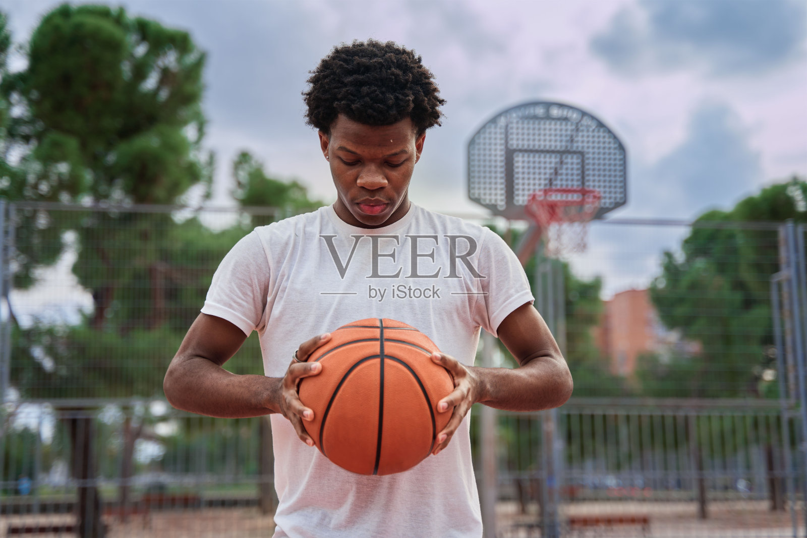 年轻英俊的非裔美国男子篮球运动员在户外双手持球。有篮球场的城市背景。男篮球运动员的肖像。照片摄影图片