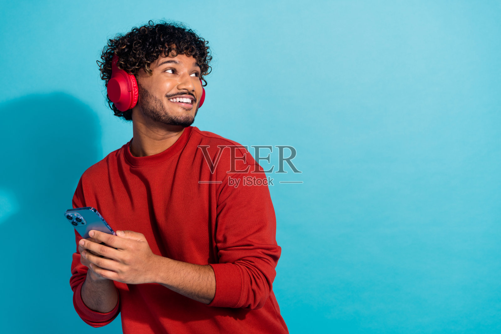 积极的美国人的肖像拿着智能手机，听音乐，耳机看空空间孤立的蓝色背景照片摄影图片