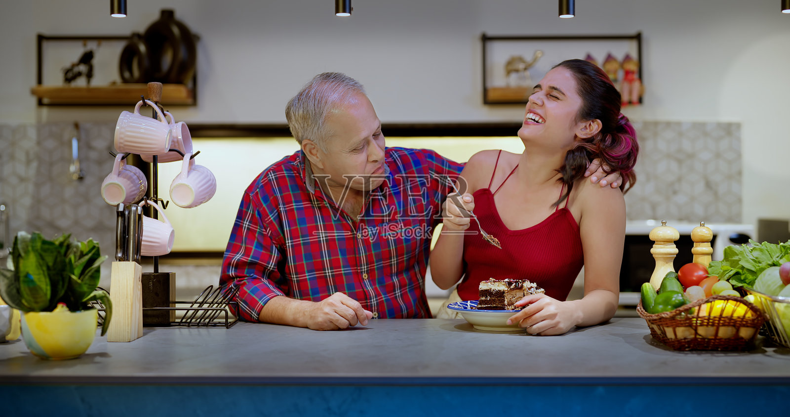 印度印度教快乐的爷爷和女儿站在现代厨房在室内的房子一起吃一块糕点。微笑的爸爸漂亮的女孩享受暑假假期，在家里玩得开心照片摄影图片