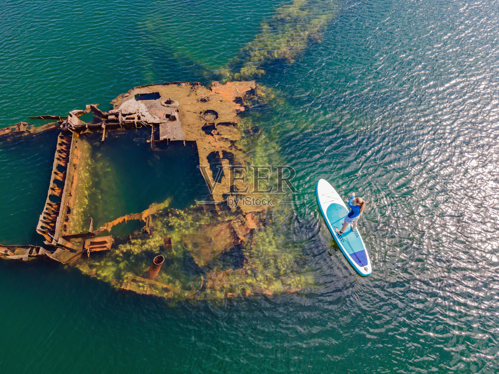 一个女人坐在划桨板上，旁边是一艘被遗弃的破船，露出海面照片摄影图片