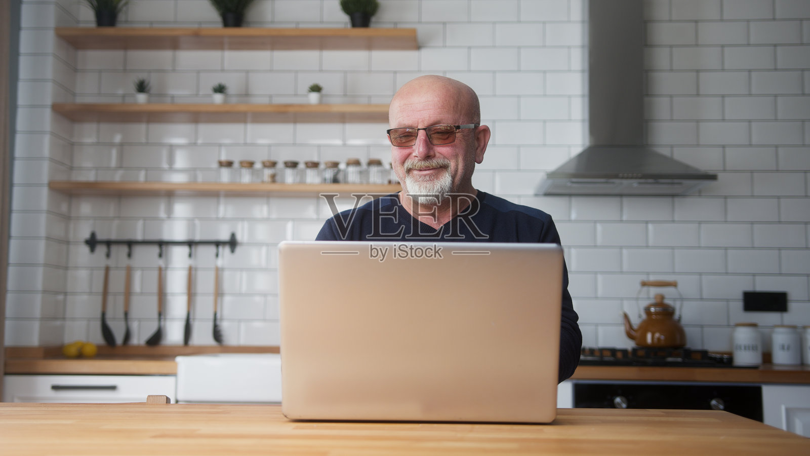 面带微笑的成熟男人戴着眼镜坐在厨房里用笔记本电脑上网，在社交媒体上享受交流，在家休息。无线，现代科技，电子购物照片摄影图片