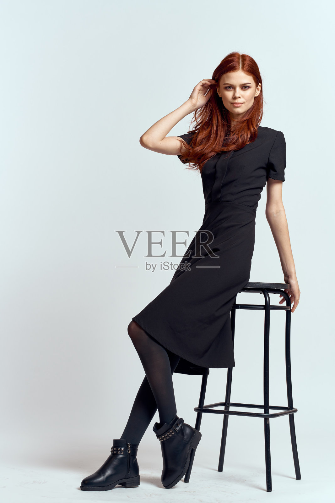 时尚的红发女人穿着黑色连衣裙坐在浅色背景的椅子上照片摄影图片