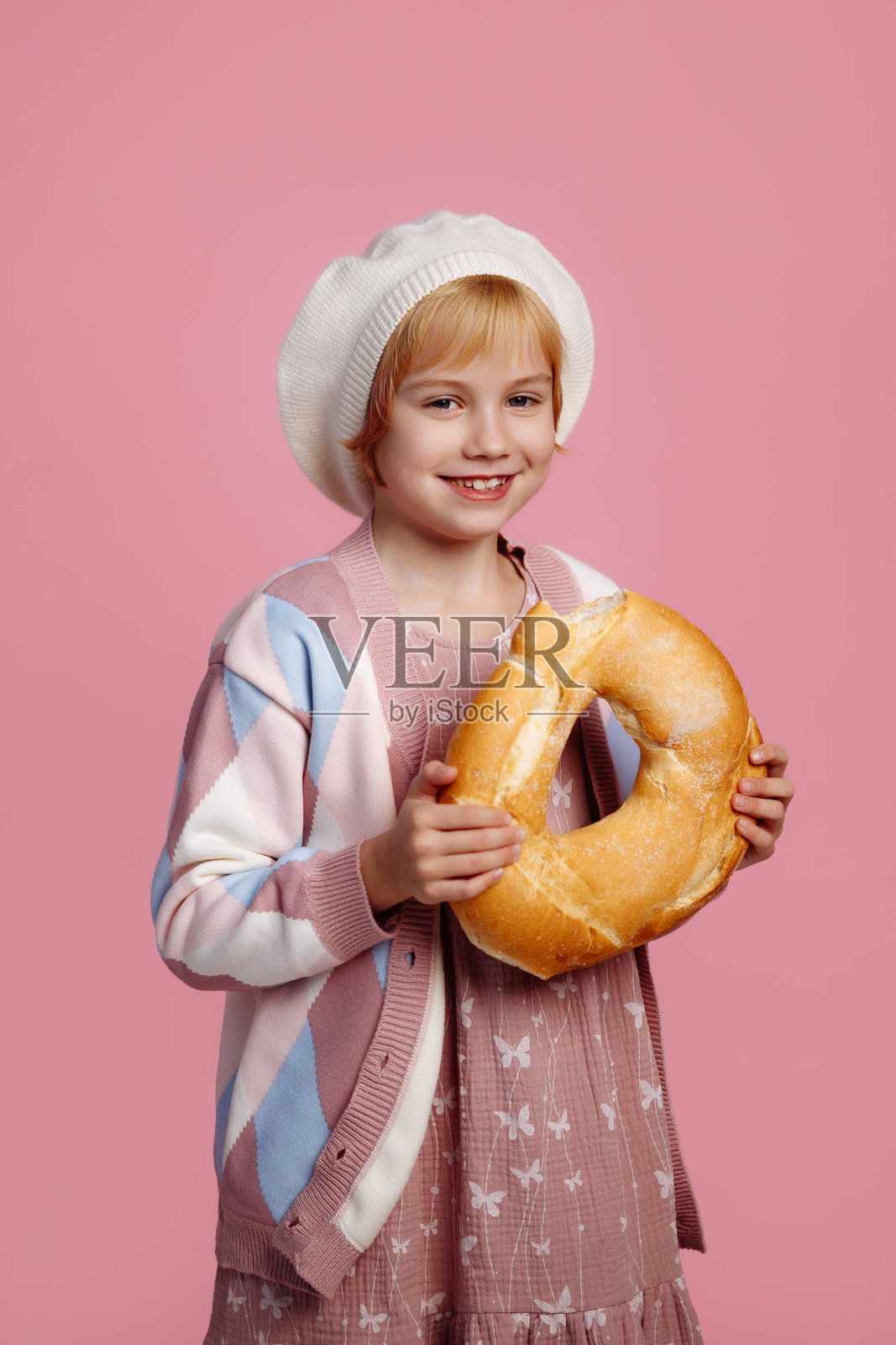 一个戴着厨师帽的孩子咬着一个大百吉饼，背景是粉红色的照片摄影图片
