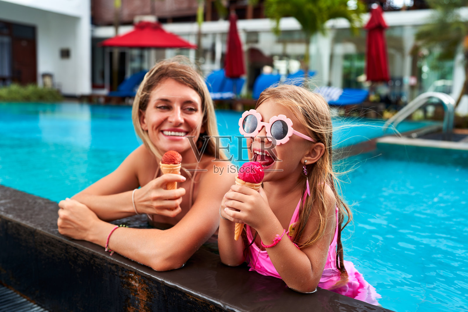 微笑的妈妈和女儿在泳池边享用冰淇淋。穿着粉色泳装，戴着花墨镜的女孩舔着蛋卷。夏天快乐的家庭时刻。度假氛围在豪华度假胜地与泳池背景。照片摄影图片