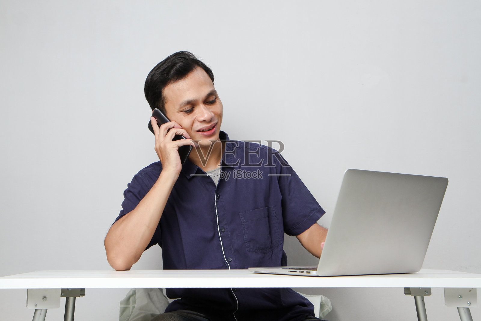 一个快乐的亚洲人坐在笔记本电脑前打电话。在孤立的背景下照片摄影图片