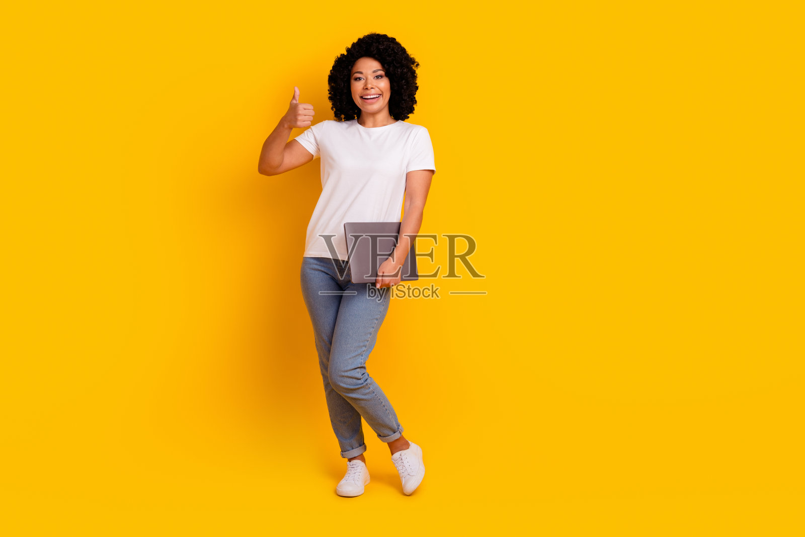 兴奋自信的女士穿着白色t恤的全身照片，拇指向上拿着现代小工具孤立的黄色背景照片摄影图片