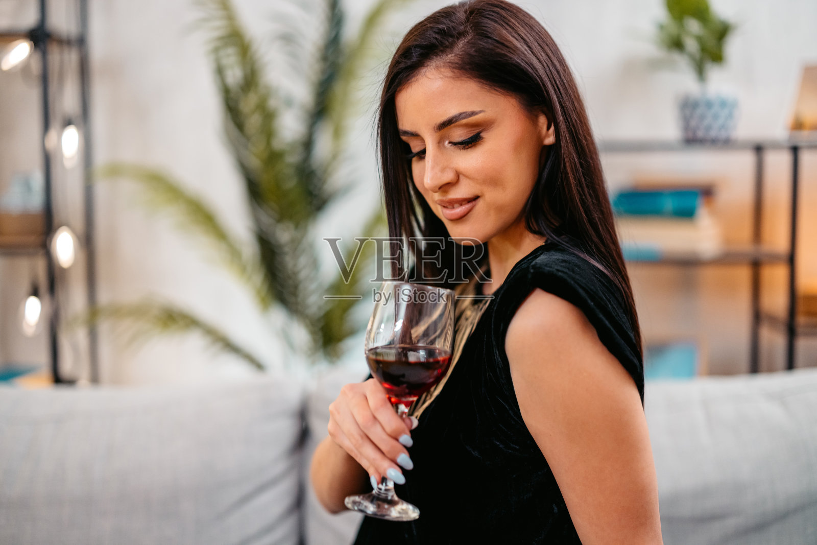 晚上，穿着裙子的年轻女子坐在客厅的沙发上喝着红酒照片摄影图片