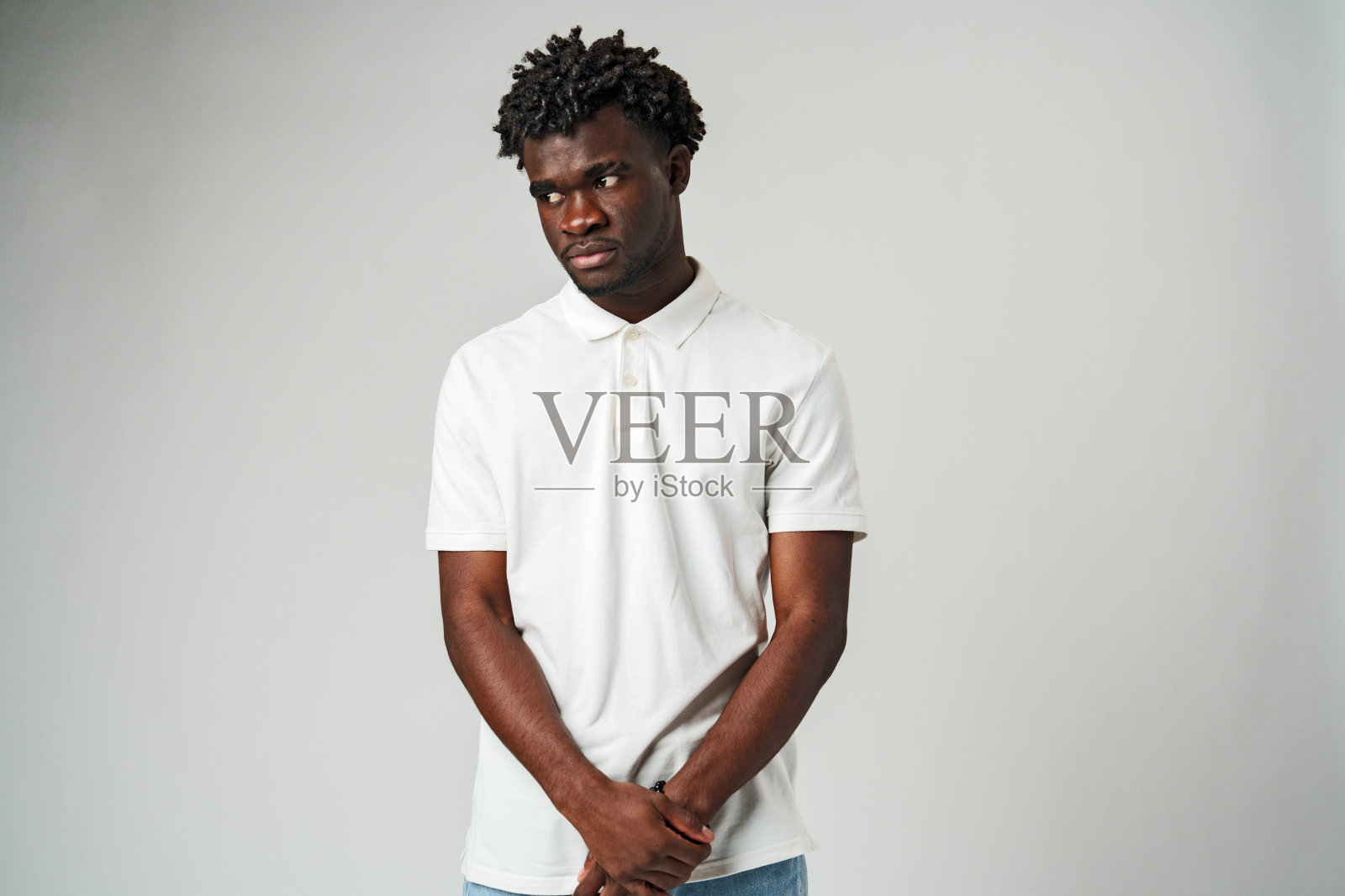 一头卷发的年轻人穿着白色Polo衫和蓝色牛仔裤站在灰色的背景下照片摄影图片