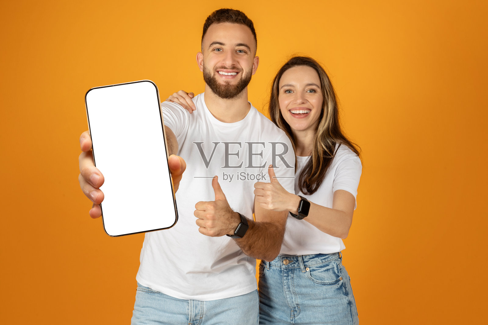 一对兴高采烈的年轻男女竖起大拇指，展示着空白屏幕的智能手机照片摄影图片