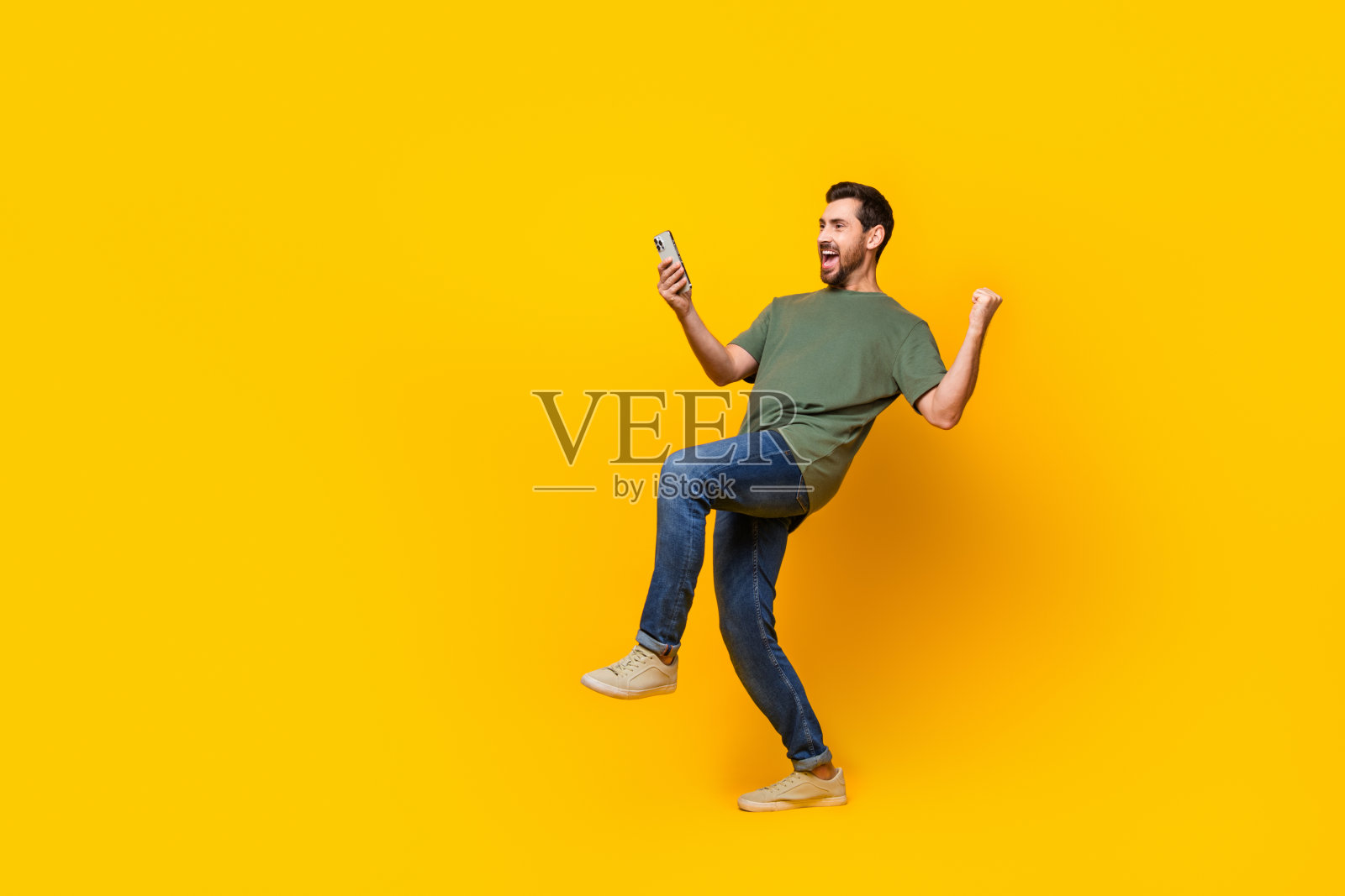 漂亮的年轻男性的全身照片，手持设备举起拳头，兴奋地穿着时尚的卡其色服装，孤立在黄色背景上照片摄影图片