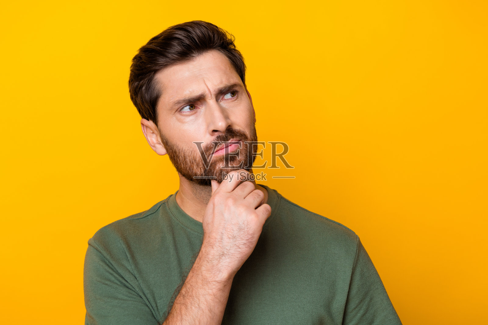 照片中若有所思的疑神疑鬼的家伙穿着绿色的t恤，手拿下巴，看起来空无一物，孤立的黄色背景照片摄影图片
