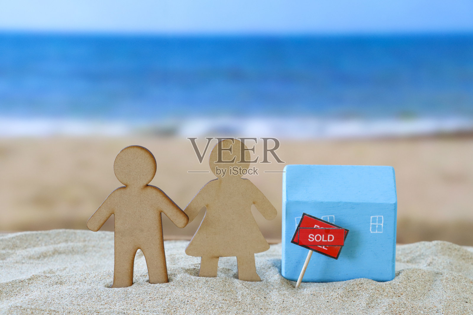 木制的异性恋夫妇的特写图像，浅蓝色，木制模型度假小屋，“出售”的标志与对角线旁边的“出售”横幅在一堆沙子上，沙滩背景与海浪在海上低潮时破碎，度假屋照片摄影图片