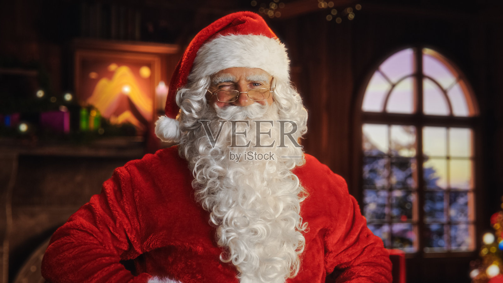 圣诞老人的肖像站在一个节日舒适的家壁炉，装饰，雪落在外面，圣诞树。圣诞老人看着镜头，微笑，眨眼。寒假庆祝活动照片摄影图片