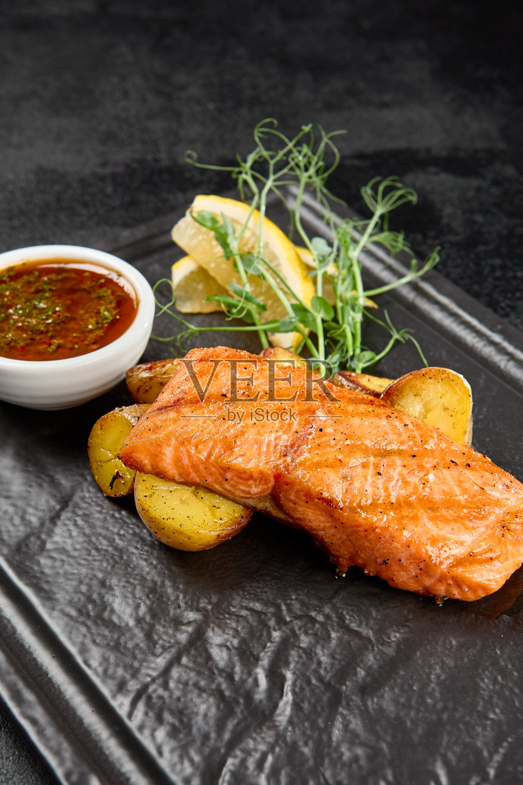 三文鱼牛排配烤土豆和酱汁，黑色石盘。三文鱼片烤在烤架上与蔬菜装饰在黑暗的背景在最小的风格。菜单上有土豆烤鲑鱼。照片摄影图片