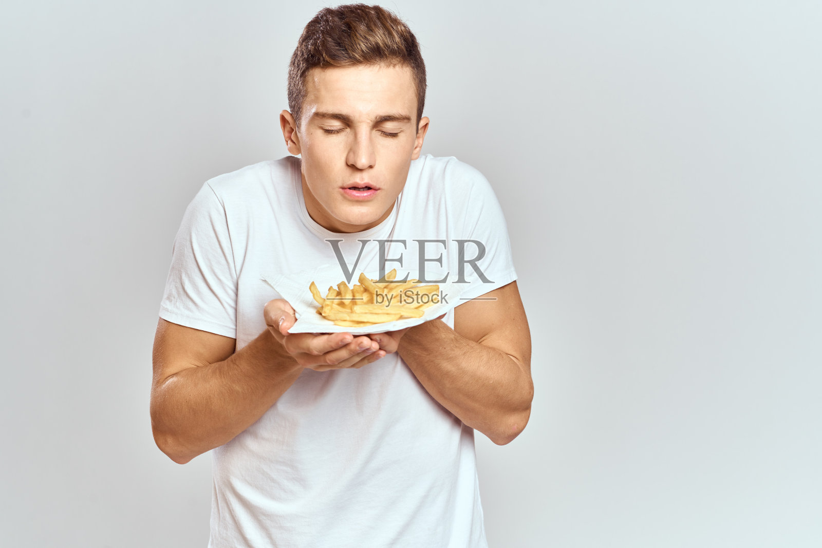 一个穿着白色t恤的年轻人拿着薯条装在盒子里，背景很淡照片摄影图片