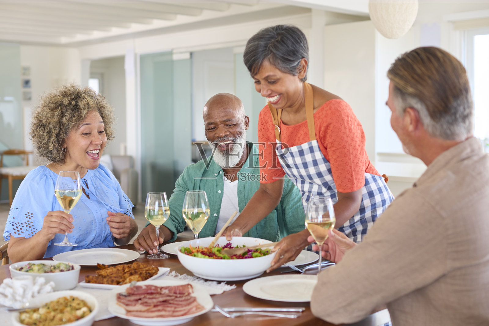 一群面带微笑的成熟朋友在家里放松地共进午餐，一起喝葡萄酒，由女士服务照片摄影图片