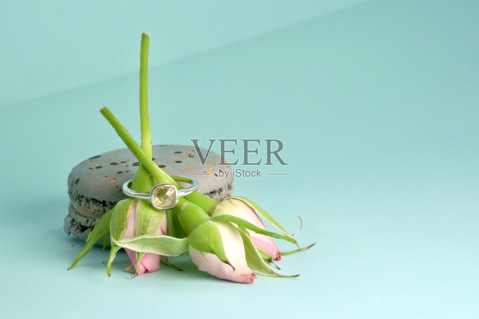 一枚镶有宝石的订婚银戒指，戴在三束粉色玫瑰花蕾上，斜倚在蓝色背景上的灰色斑点通心粉蛋糕上照片摄影图片