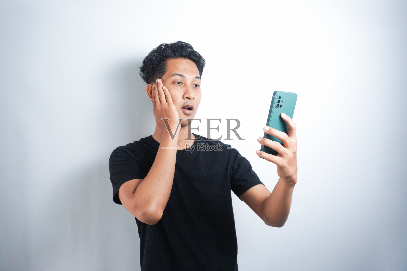 哇，你的亚洲男人的脸震惊了他在智能手机上看到的孤立的灰色背景。印尼男子身穿黑色衬衫，背景为灰色。照片摄影图片